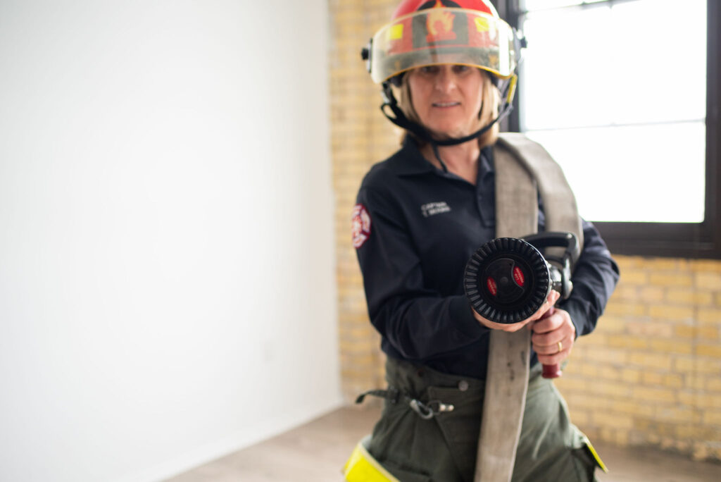 Fire captain Tracy Moore holds a fire hose nozzle facing the camera. She has red helmet and a flame on the front of the helmet.